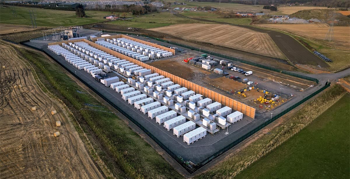 Aerial view of Zenobē’s Blackhillock battery storage site, showcasing rows of white battery containers, wooden wind breaker panels, and surrounding green fields.