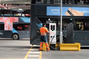Man in orange hi-viz tending to a Zenobe branded EV charger next to a National Express electric bus